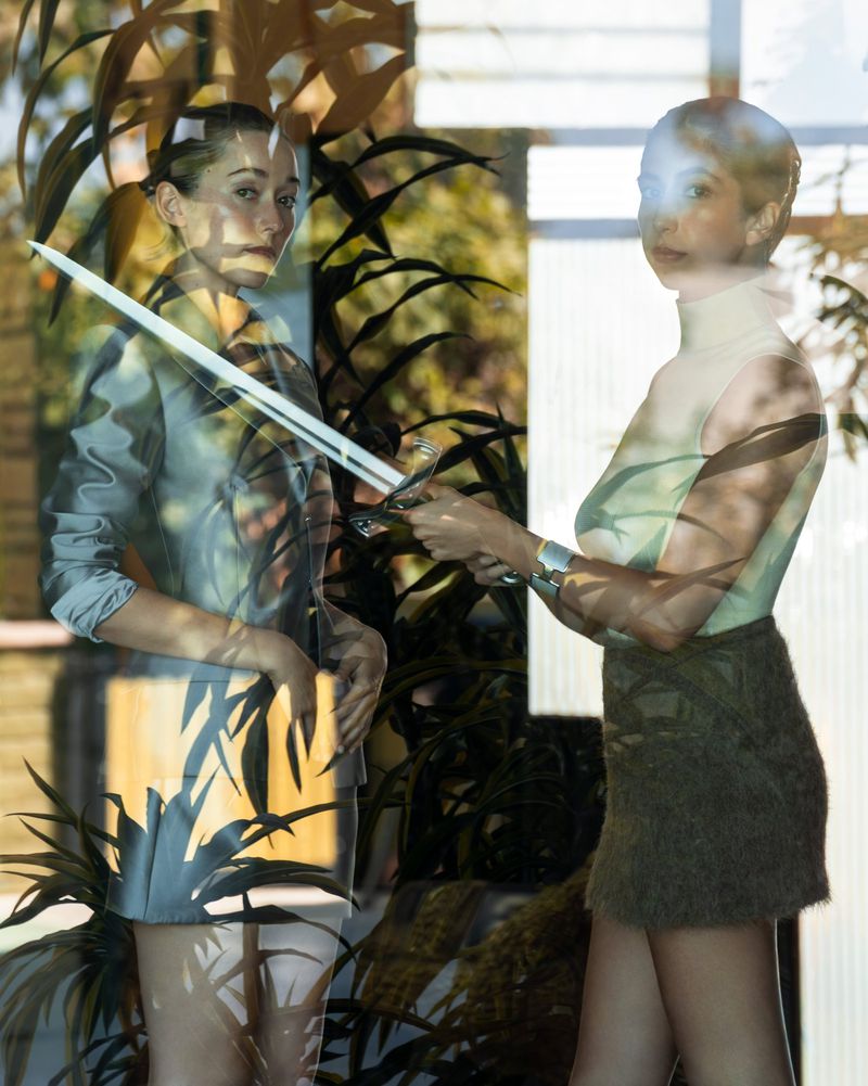 Two women posing indoors with foliage reflections layered across the glass, holding a long sword together.