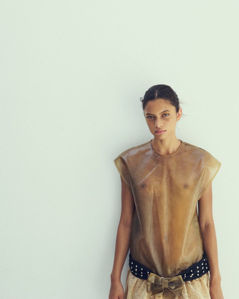 Model wearing a sheer brown top and a dark studded belt standing against a plain white wall