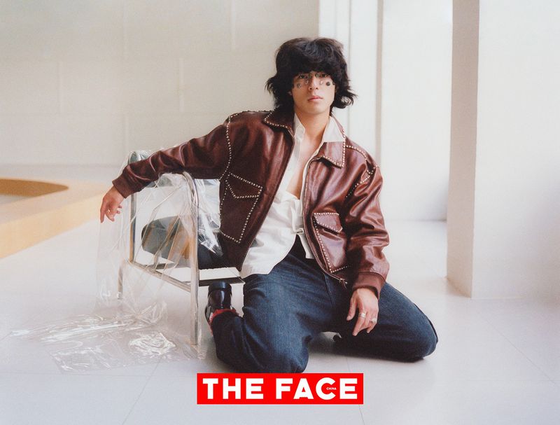 A man with dark hair kneeling in a brown leather jacket and jeans on a white floor next to a transparent plastic-wrapped chair