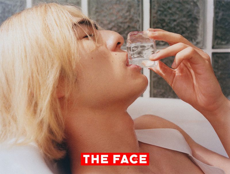 Close-up portrait of a blonde man holding an ice cube to his lips against a textured glass tile background.
