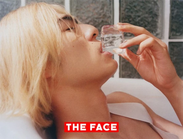 Close-up portrait of a blonde man holding an ice cube to his lips against a textured glass tile background.