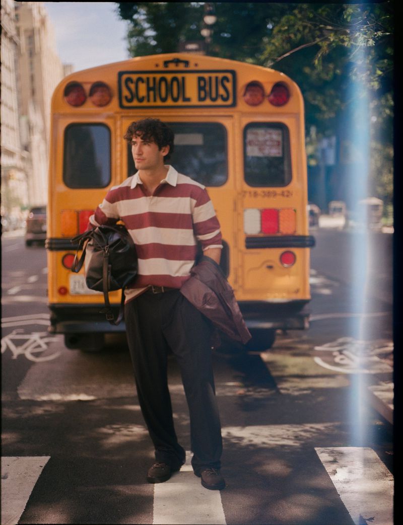 Man standing on a street in front of a yellow school bus wearing a striped polo shirt, dark trousers, and carrying a duffel bag.