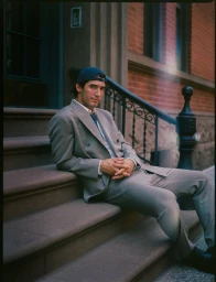 Man wearing a grey double breasted suit and blue baseball cap seated on brown stone steps outdoors