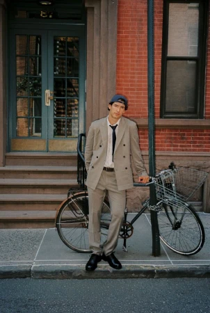 Man in a tan double-breasted suit leaning against a bicycle in front of a red brick building with teal double doors.