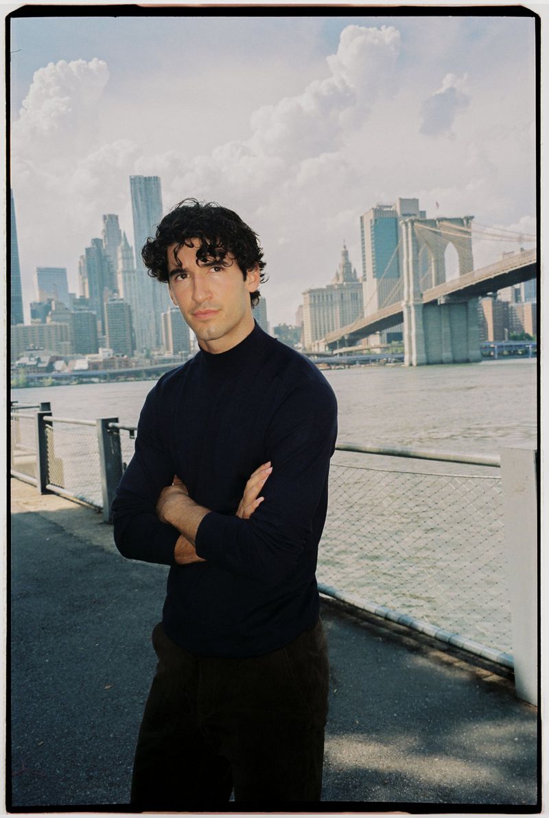 A man in a dark sweater stands with arms crossed outdoors by a waterfront with a bridge and city skyline background.