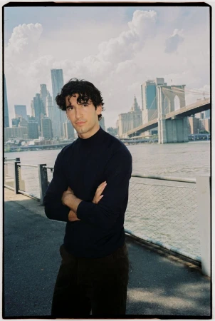 A man in a dark sweater stands with arms crossed outdoors by a waterfront with a bridge and city skyline background.