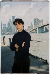 A man in a dark sweater stands with arms crossed outdoors by a waterfront with a bridge and city skyline background.