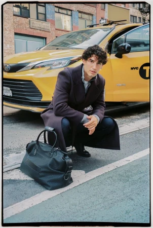 Man crouching beside a yellow taxi cab wearing a dark coat with a black duffel bag on the asphalt street.