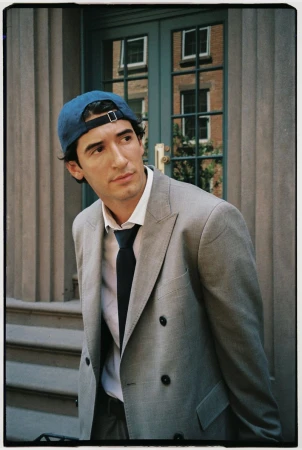 Man wearing a grey double-breasted suit jacket, white shirt, dark tie, and blue baseball cap looking away.