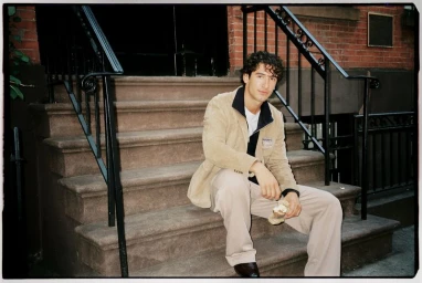 A man in a tan jacket sitting on brownstone steps holding food, photographed for the Reserved men's fall campaign.