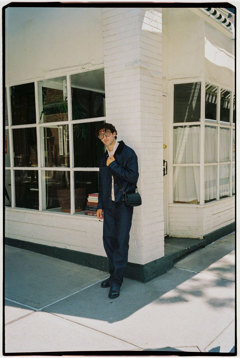 Man wearing a dark blue jacket and trousers leaning against a white brick building exterior holding a bag