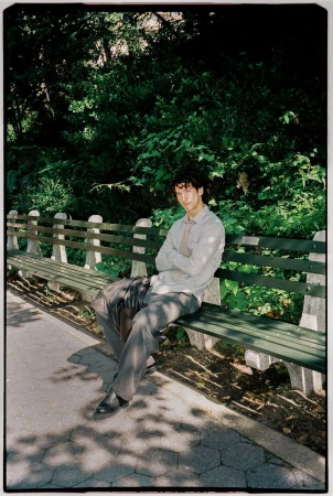 A man sitting on a park bench with his arms crossed, wearing a light shirt and dark trousers.