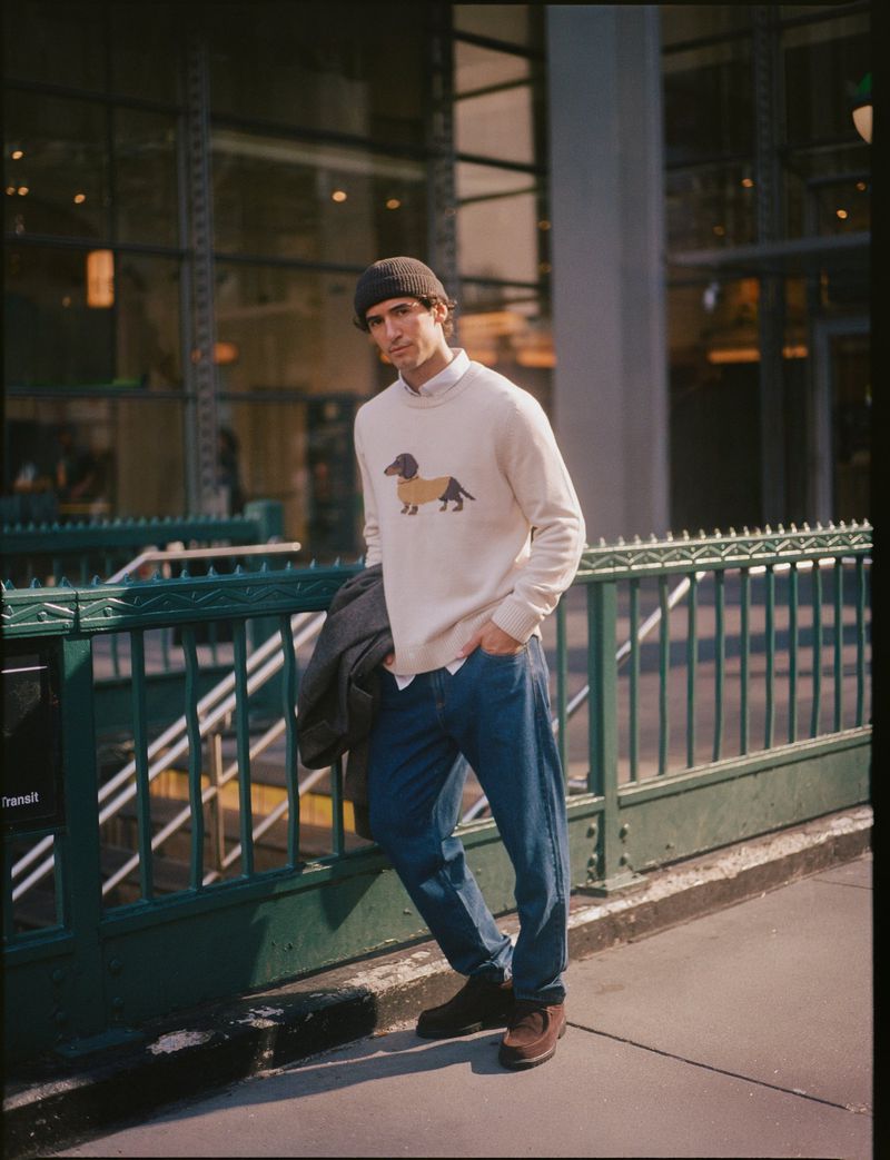 Man wearing white dachshund-intarsia sweater, dark beanie, and blue jeans leaning against a green transit railing outdoors