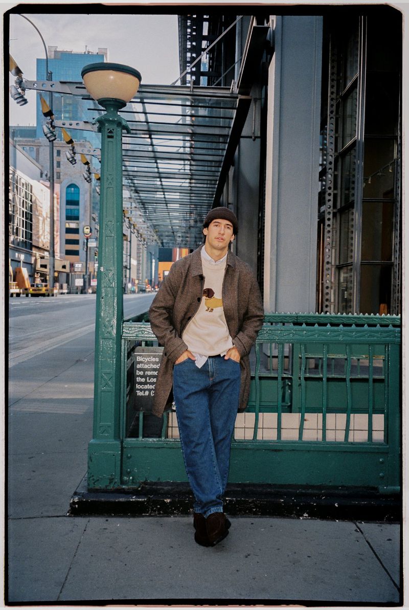 Man wearing a brown wool coat, beanie, and jeans posing on a city sidewalk next to a green subway entrance.
