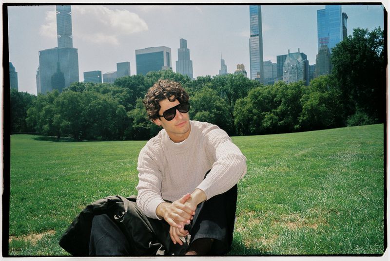 Man wearing sunglasses and a textured beige sweater sitting on a grassy park lawn with city skyline