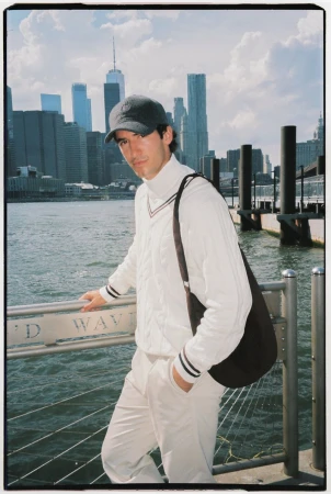 Man wearing a white sweater, cap, and carrying a shoulder bag leaning against a waterfront railing with city skyline behind.