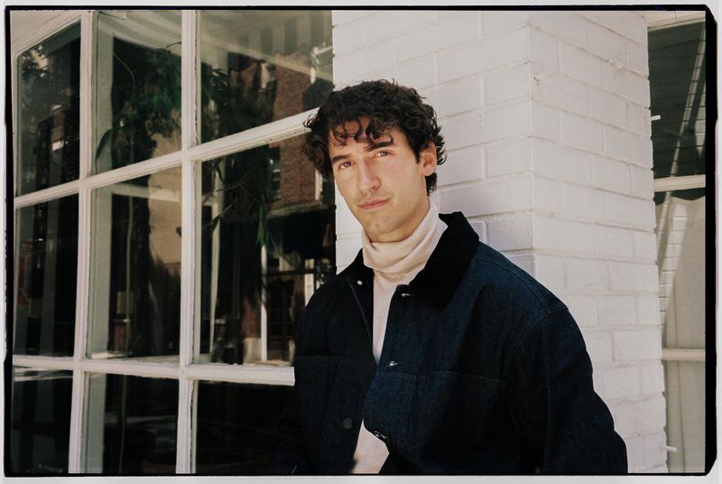 A man with dark curly hair wearing a dark jacket and turtleneck stands in front of a white brick column and large window.