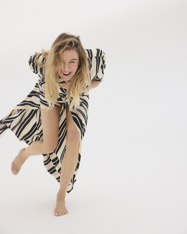 A blonde woman in a striped dress strikes a playful pose against a bright white backdrop, showcasing a blend of fashion and fun.