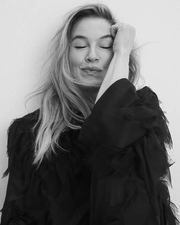 Black and white portrait of a blonde woman with eyes closed and hand on her head wearing a black top against a neutral backdrop.