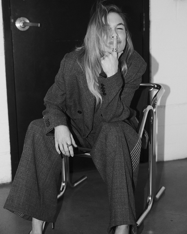 Black and white portrait of Renee Zellweger seated in a chrome chair wearing a tailored tweed suit