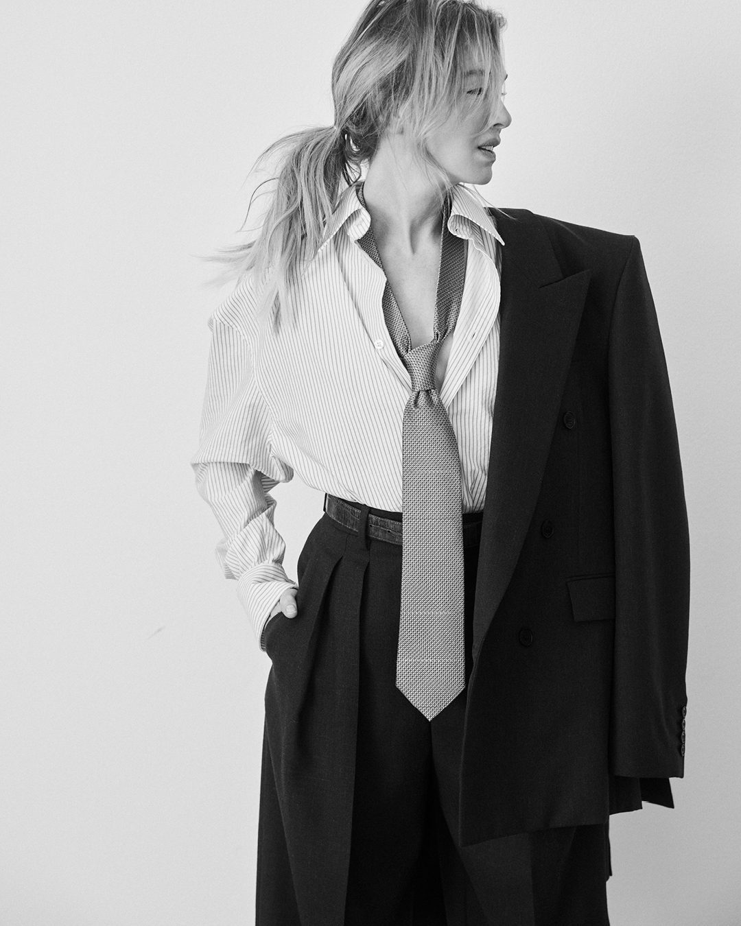 Black and white portrait of a blonde woman in pinstripe shirt, tie, and blazer draped over her shoulder, standing indoors.