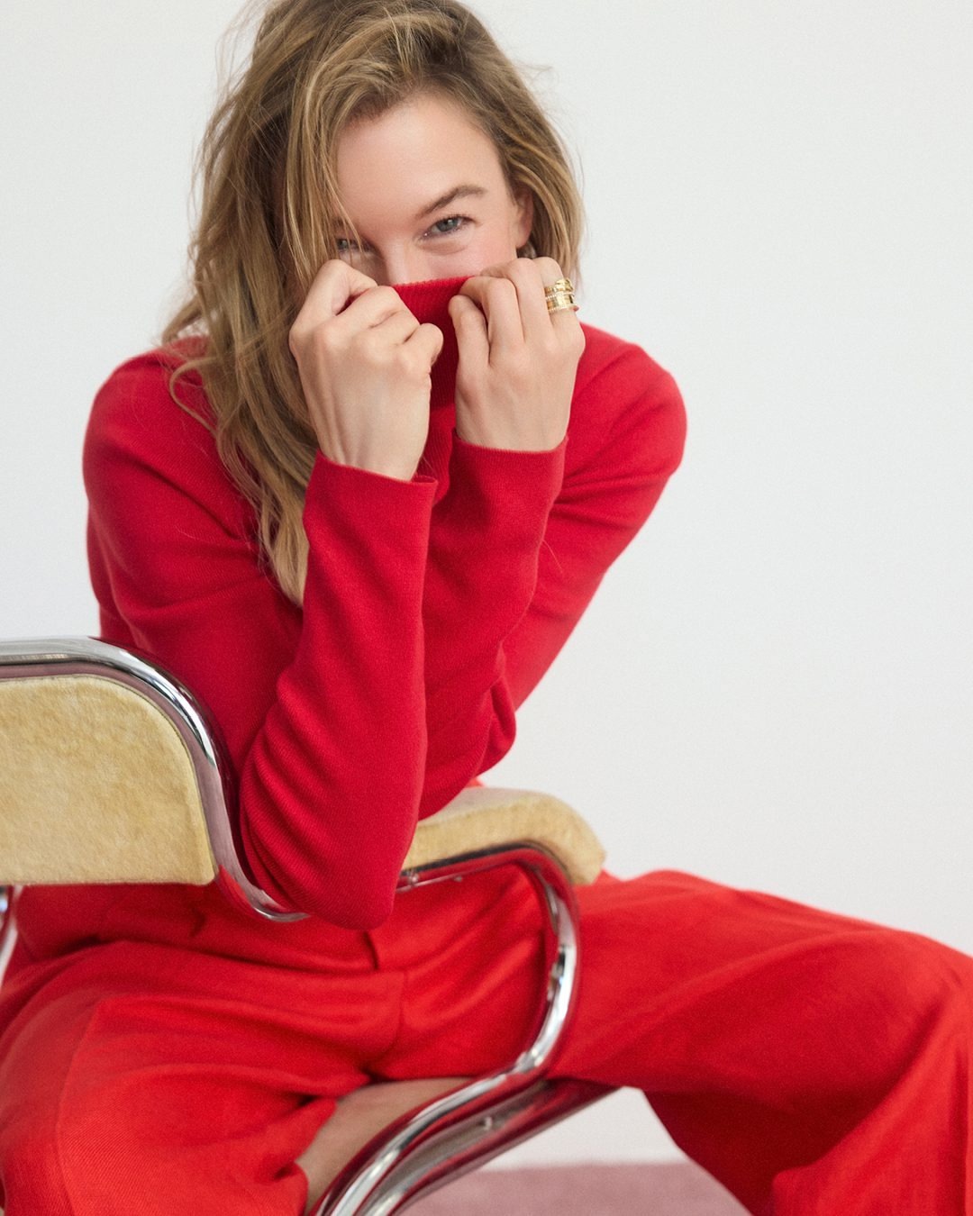 Renee Zellweger wearing a red turtleneck sweater and trousers, holding the collar up to her face against a white seamless background