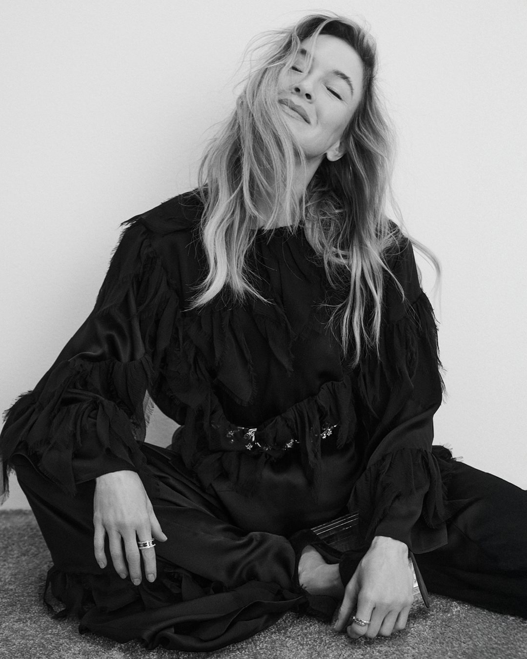 Black and white portrait of a middle-aged woman with blonde wavy hair, wearing a black outfit while sitting in a photo studio.