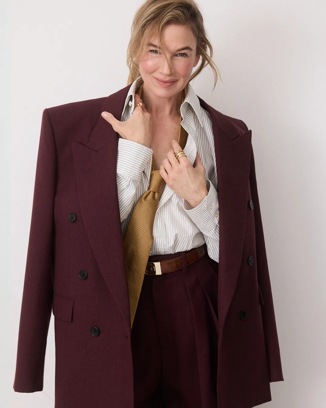Renee Zellweger wearing a burgundy blazer over a striped shirt and gold tie against a plain white wall