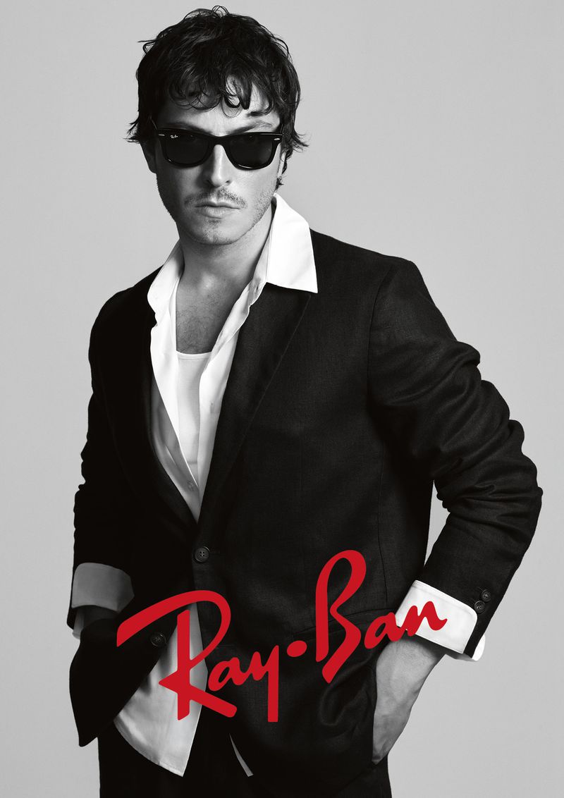 Boran Kuzum wearing Ray-Ban sunglasses and a black suit jacket over a white collared shirt against a light grey background.