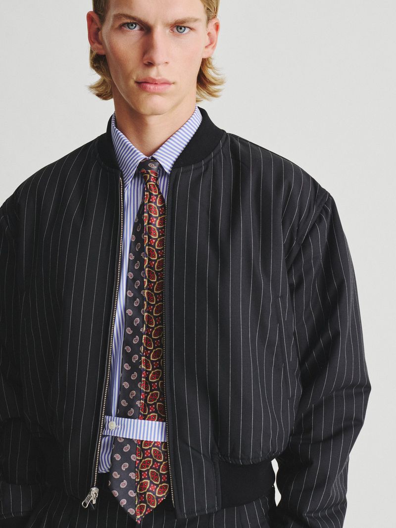 Model wearing a black pinstriped bomber jacket over a striped shirt and patterned tie against a light background.