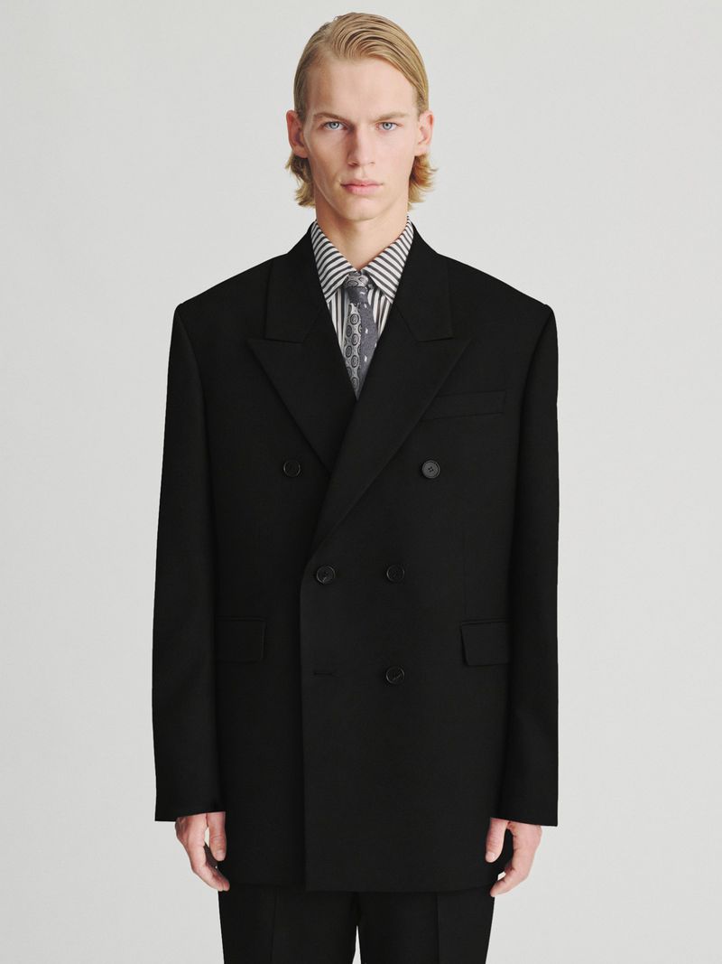 Model in black double-breasted suit jacket with striped shirt and tie against a plain white background