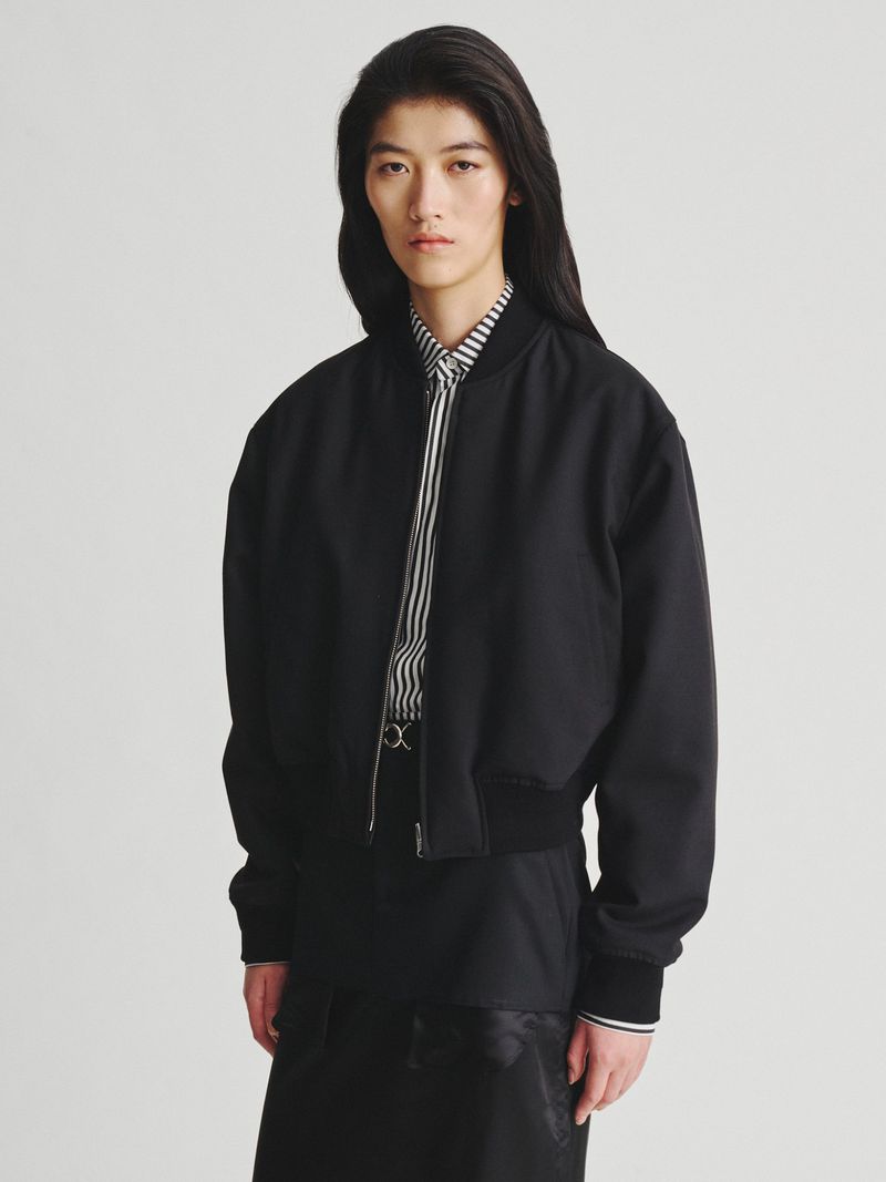 Model with long dark hair wearing a black bomber jacket over a striped shirt against a plain white background