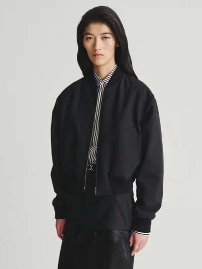 Model with long dark hair wearing a black bomber jacket over a striped shirt against a plain white background