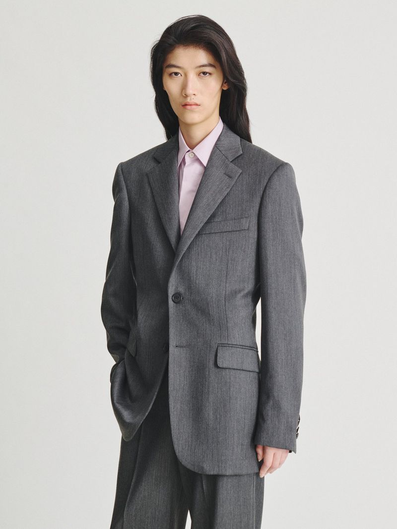 Model with long dark hair wearing a charcoal grey suit jacket over a pink button-down shirt against a white backdrop.