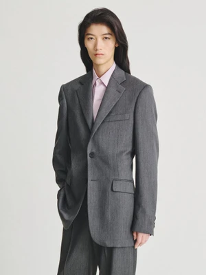 Model with long dark hair wearing a charcoal grey suit jacket over a pink button-down shirt against a white backdrop.