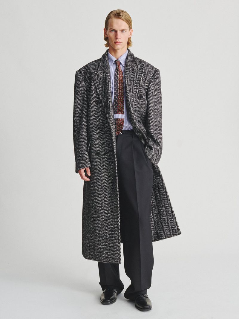Male model in an oversized grey herringbone trench coat, patterned tie, dark trousers, and leather shoes against a white background