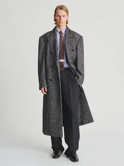 Male model in an oversized grey herringbone trench coat, patterned tie, dark trousers, and leather shoes against a white background