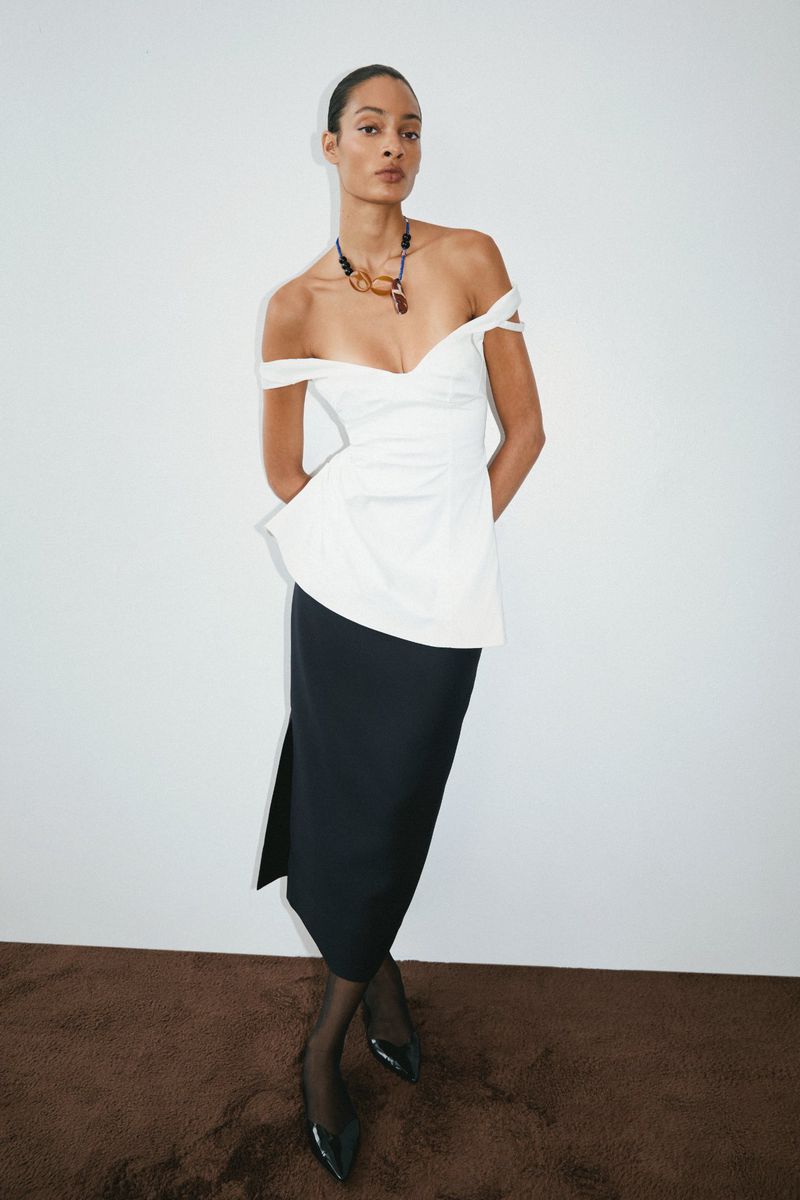 Model in off-the-shoulder white top and black skirt wearing a beaded necklace against a plain white background