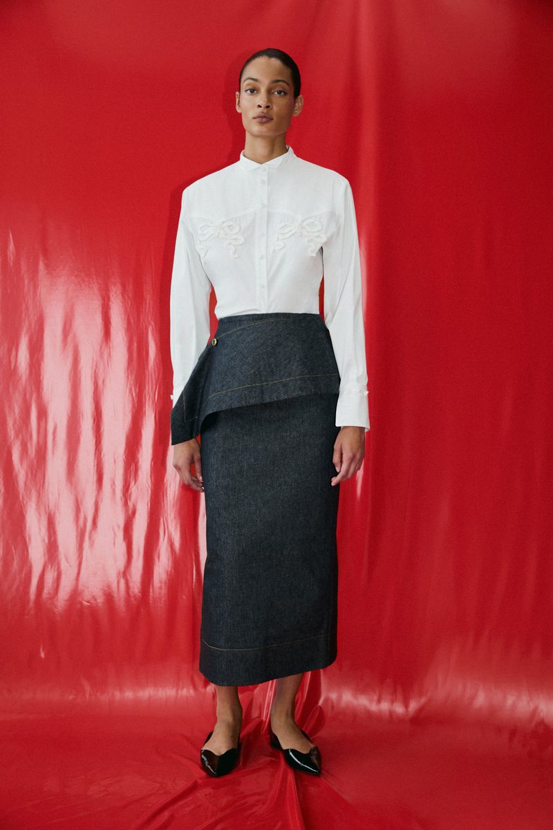 Model standing against a vibrant red backdrop wearing a white long-sleeve button-down shirt and a dark denim midi skirt with a draped peplum overlay.