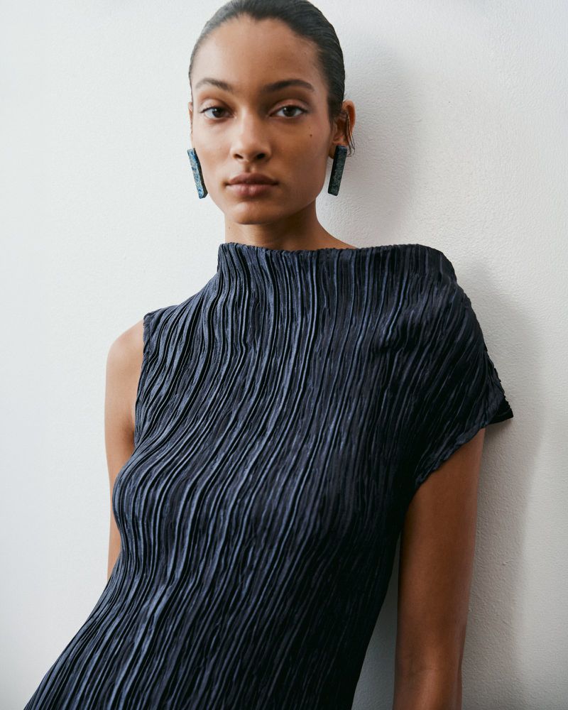 Model wearing a pleated dark top and statement earrings against a plain white wall for Amir Taghi