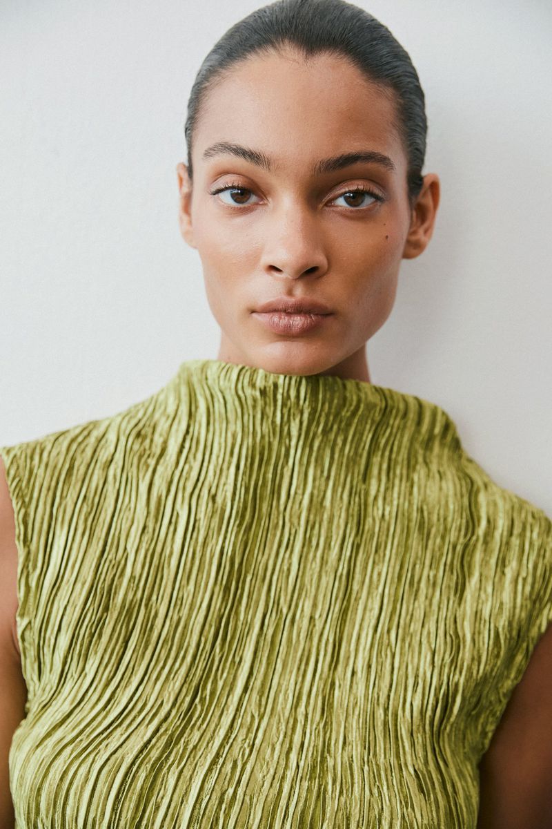Close-up portrait of a model wearing a textured chartreuse pleated sleeveless top against a white wall.