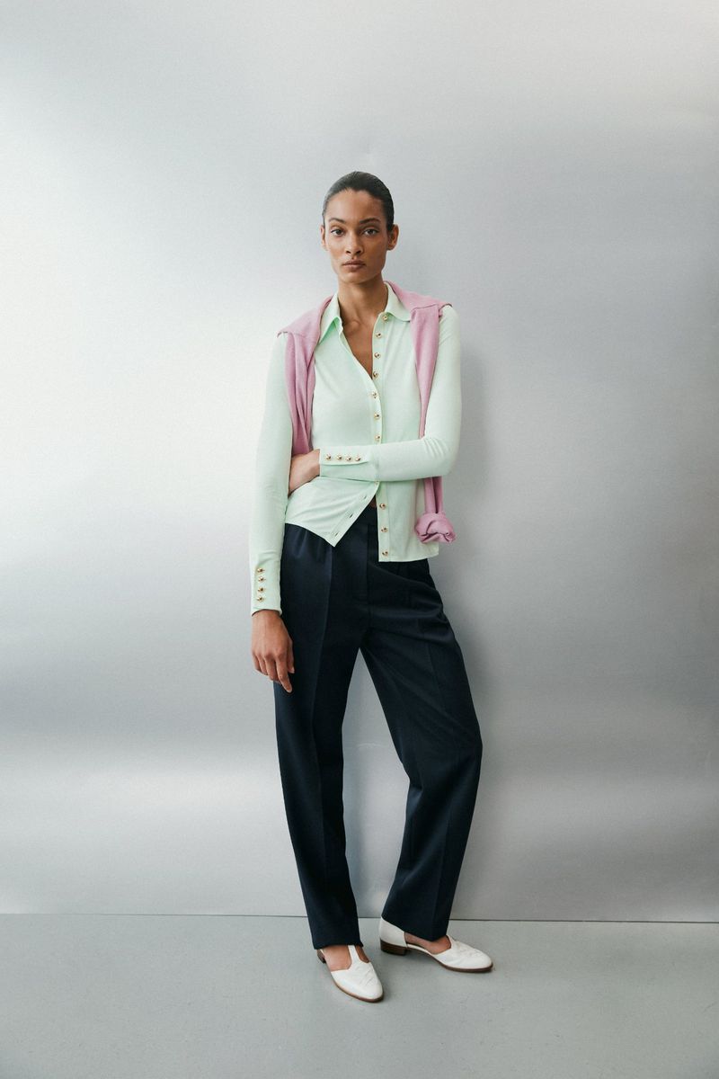 Model standing against a grey seamless backdrop wearing a light green buttoned top, dark trousers, and white shoes