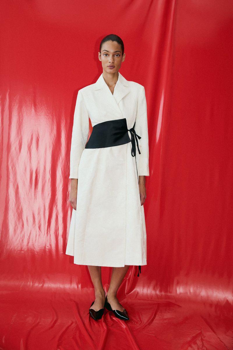 Model in a white midi wrap coat with a wide black waist tie standing against a glossy red backdrop