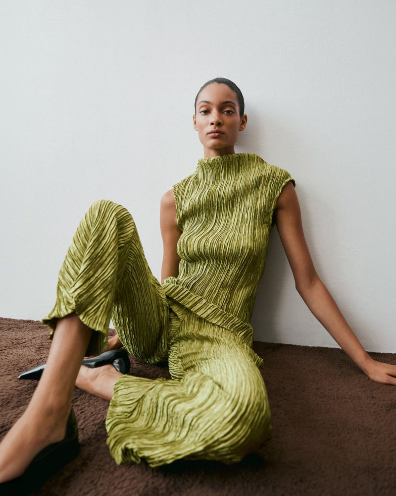 Model wearing a pleated olive green top and matching trousers sitting on a brown carpet against a white wall