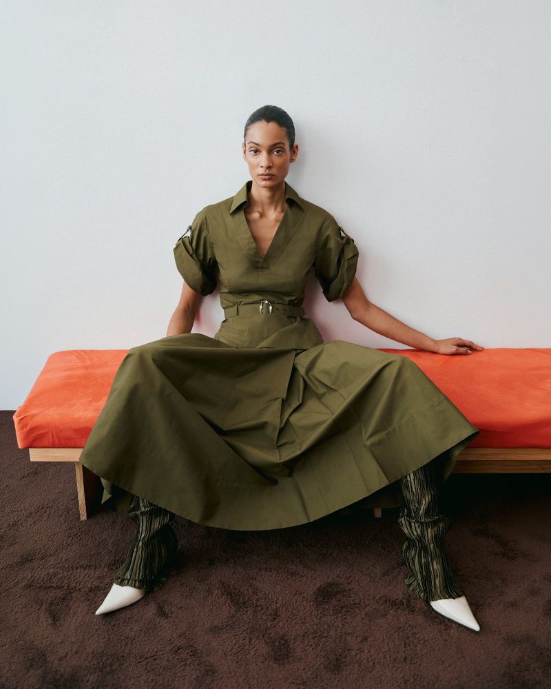 Model wearing an olive green dress and white pointy heels seated on an orange bench against a plain white wall