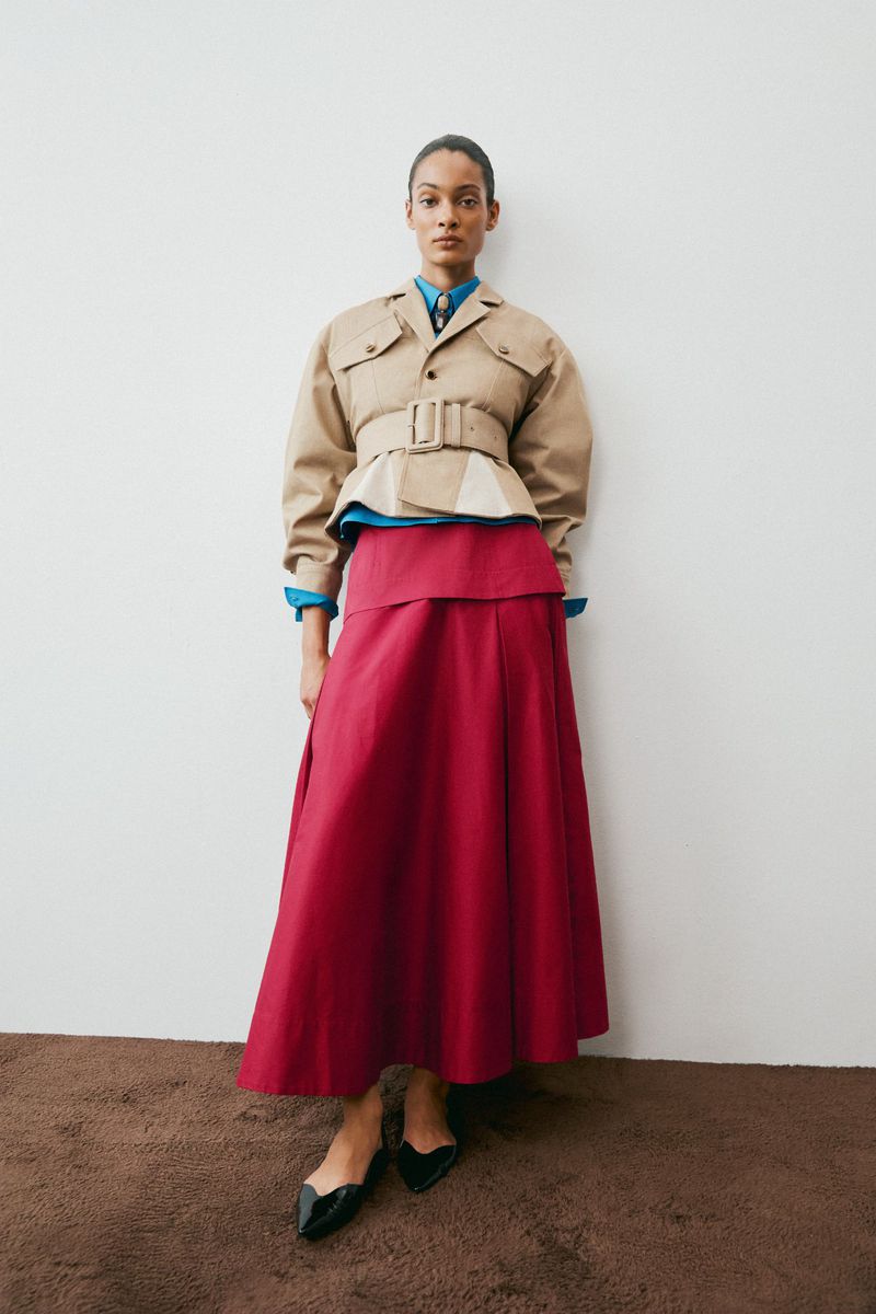 Model in a belted khaki jacket and bright pink midi skirt standing against a white backdrop.