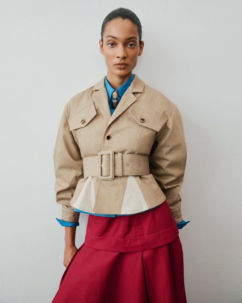 Model wearing a tan belted jacket over a bright blue shirt and crimson skirt against a plain white background
