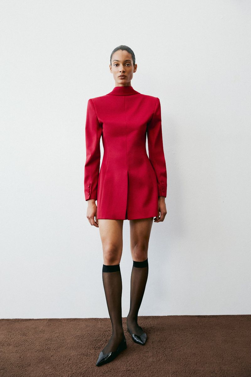 Model wearing a structured red long-sleeve mini dress and sheer black knee-high socks against a white backdrop.