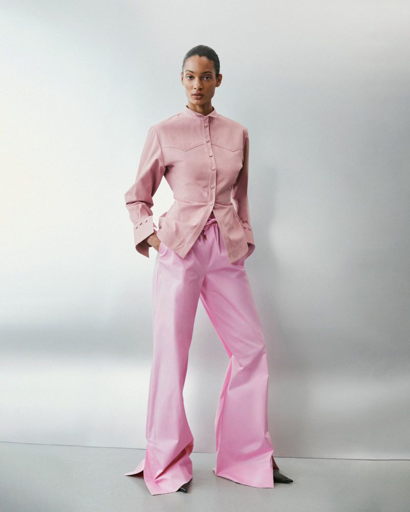 Model wearing a pink button-down shirt and matching trousers against a pale grey seamless studio backdrop