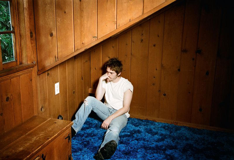 Actor Austin Abrams seated on a blue carpet in a wood-paneled room wearing a white t-shirt and light blue jeans